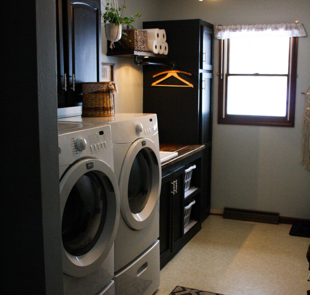 Inspiration for a mid-sized industrial single-wall dedicated laundry room in Other with an utility sink, shaker cabinets, black cabinets, wood benchtops, blue walls, linoleum floors, a side-by-side washer and dryer, beige floor and brown benchtop.