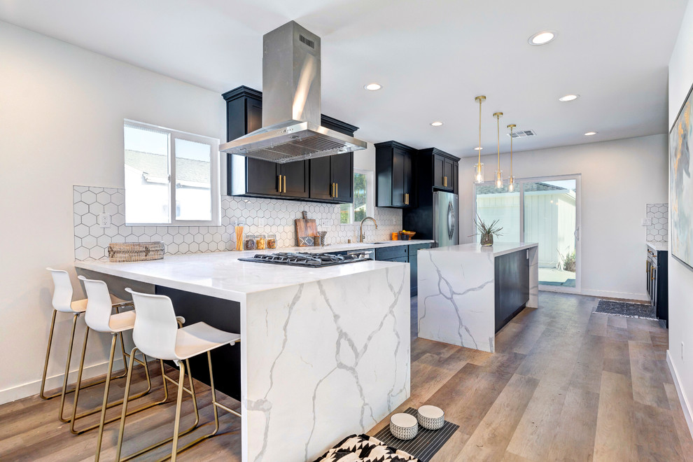 Photo of a transitional l-shaped kitchen in Los Angeles with an undermount sink, shaker cabinets, black cabinets, white splashback, stainless steel appliances, medium hardwood floors, with island, brown floor and white benchtop.