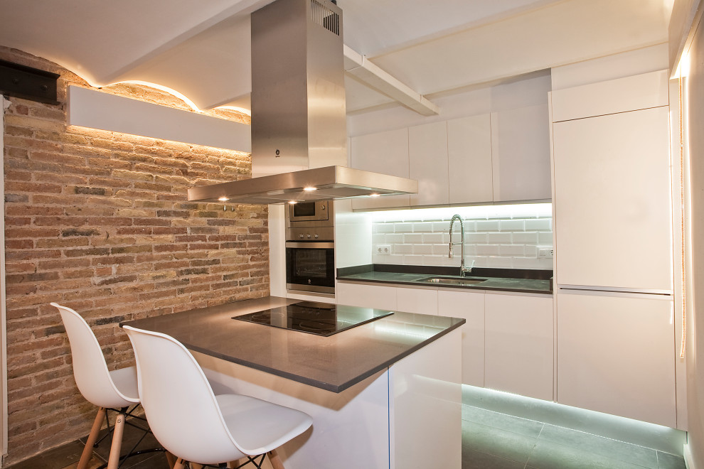 This is an example of a small industrial galley kitchen in Barcelona with an undermount sink, flat-panel cabinets, white cabinets, white splashback, porcelain splashback, panelled appliances, with island, grey floor and black benchtop.