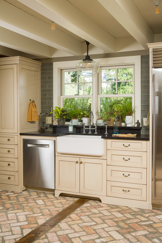 This is an example of a country kitchen in Other with a farmhouse sink, shaker cabinets, beige cabinets, stainless steel appliances and brick floors.