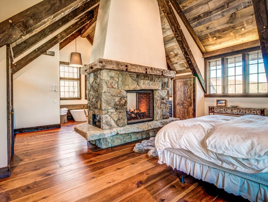 Photo of a country master bedroom in Other with dark hardwood floors, a two-sided fireplace, a stone fireplace surround, brown floor and exposed beam.