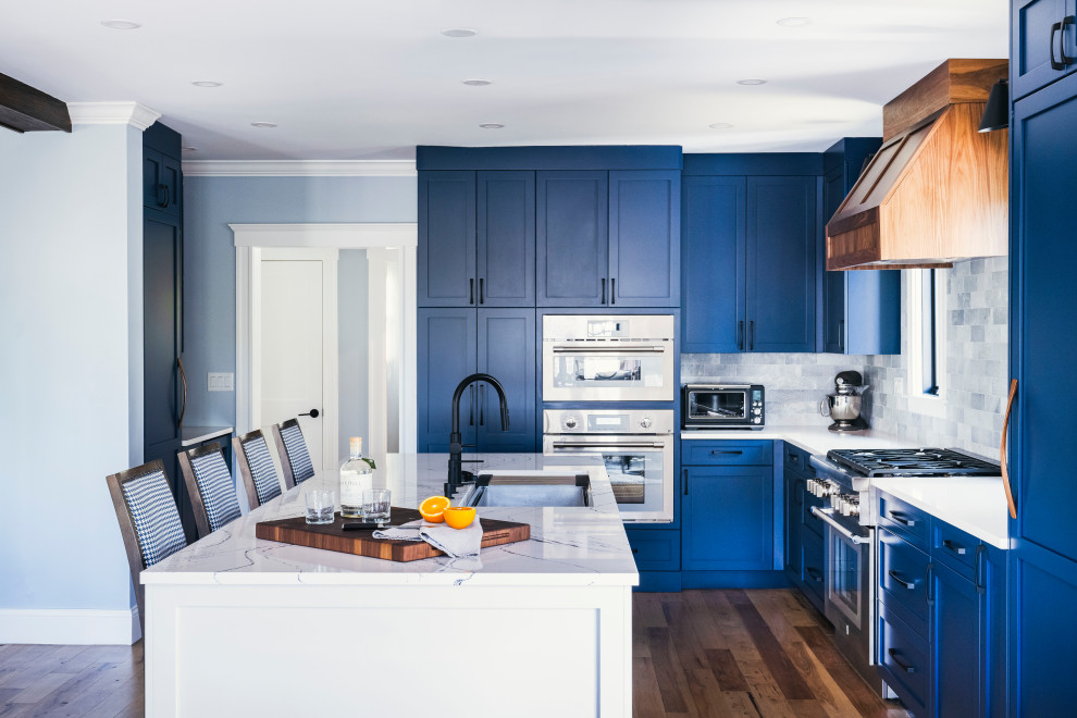 Klassisk inredning av ett vit vitt l-kök, med en undermonterad diskho, skåp i shakerstil, blå skåp, marmorbänkskiva, grått stänkskydd, stänkskydd i stenkakel, rostfria vitvaror, mellanmörkt trägolv och en köksö