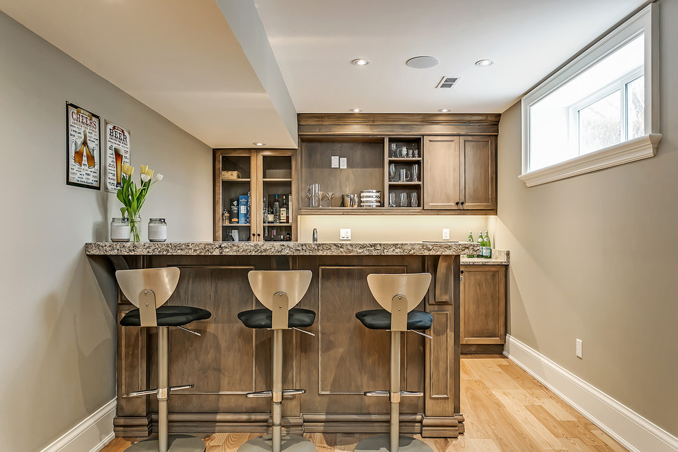 This is an example of a mid-sized transitional galley seated home bar in Toronto with an undermount sink, shaker cabinets, medium wood cabinets, granite benchtops and light hardwood floors.