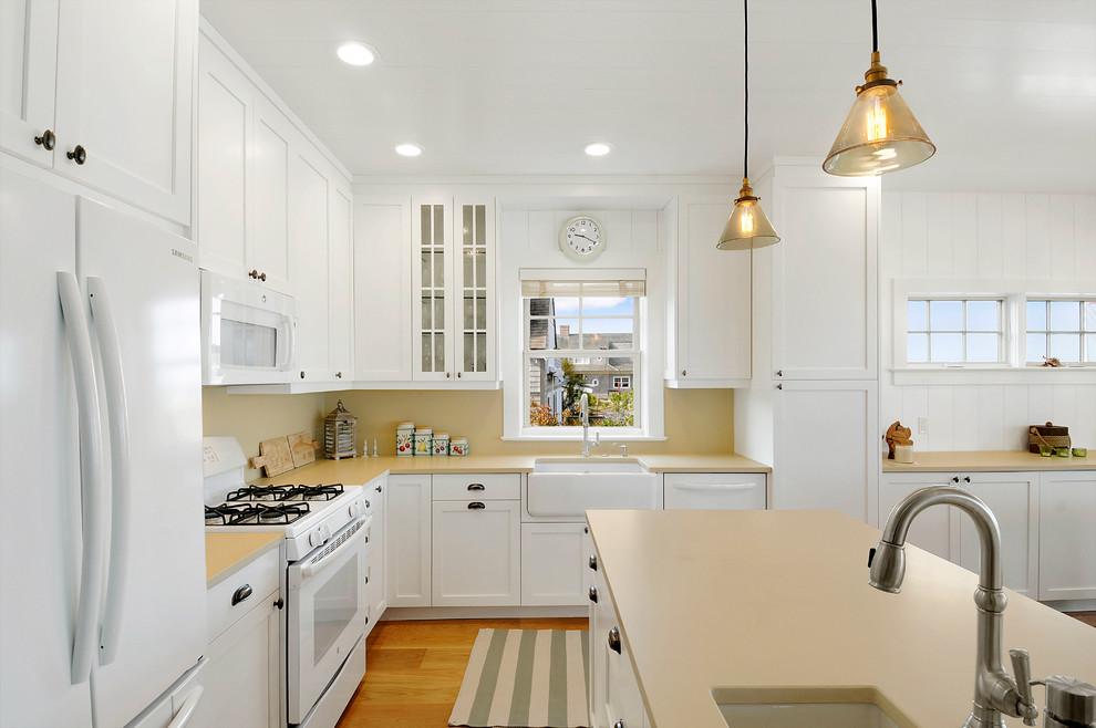 Inspiration for a small coastal l-shaped light wood floor eat-in kitchen remodel with a farmhouse sink, shaker cabinets, white cabinets, solid surface countertops, beige backsplash, white appliances and a peninsula