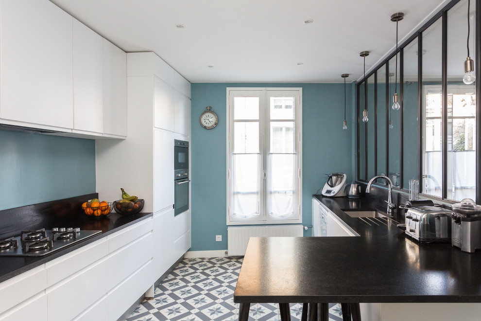 This is an example of a large contemporary galley open plan kitchen in Paris with an undermount sink, flat-panel cabinets, white cabinets, black appliances, a peninsula, grey floor, granite benchtops, black splashback, stone slab splashback, cement tiles and black benchtop.