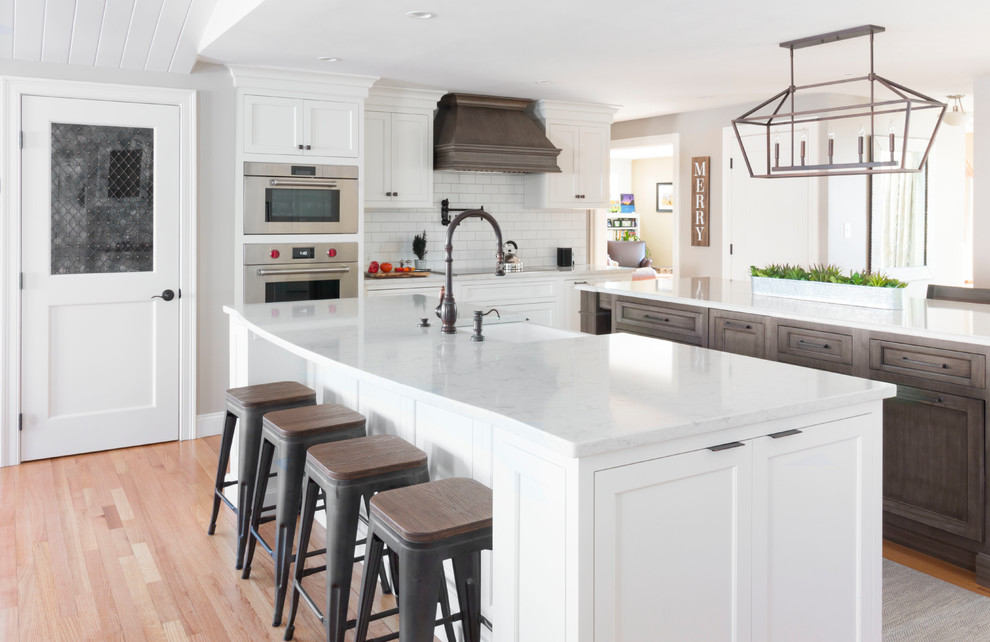 Inspiration for a country u-shaped kitchen in Boston with a farmhouse sink, shaker cabinets, white cabinets, white splashback, subway tile splashback, stainless steel appliances, light hardwood floors, multiple islands, beige floor and white benchtop.