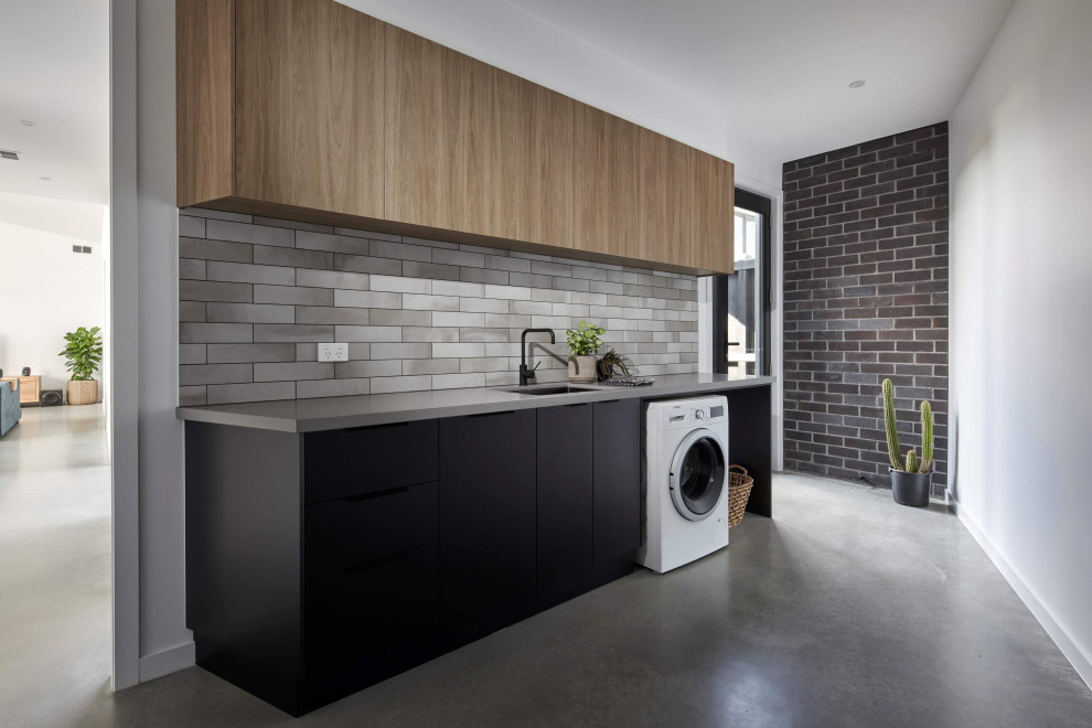Contemporary single-wall dedicated laundry room in Melbourne with a single-bowl sink, flat-panel cabinets, grey splashback, concrete floors, grey floor, grey benchtop and brick walls.