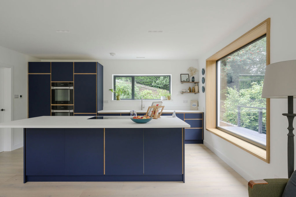This is an example of a large contemporary single-wall open plan kitchen in Surrey with a built-in sink, flat-panel cabinets, blue cabinets, composite countertops, black appliances, light hardwood flooring, an island and white worktops.