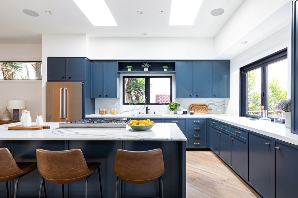 Photo of a contemporary l-shaped kitchen in Los Angeles with an undermount sink, shaker cabinets, blue cabinets, white splashback, stone slab splashback, stainless steel appliances, light hardwood floors, with island, beige floor and white benchtop.