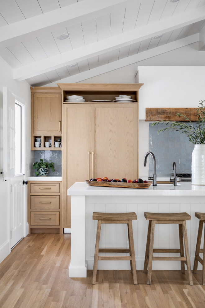 This is an example of a small coastal single-wall open plan kitchen in Orange County with shaker cabinets, light wood cabinets, engineered stone countertops, blue splashback, terracotta splashback, integrated appliances, an island, white worktops and a vaulted ceiling.