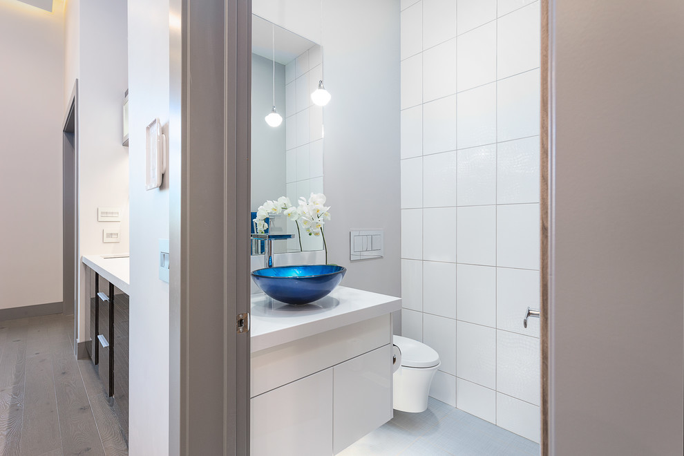 Inspiration for a mid-sized modern powder room in Edmonton with flat-panel cabinets, white cabinets, a wall-mount toilet, white tile, ceramic tile, grey walls, ceramic floors, a trough sink, engineered quartz benchtops and grey floor.