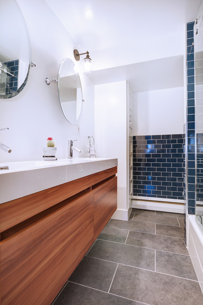 Photo of a mid-sized transitional master bathroom in Denver with flat-panel cabinets, medium wood cabinets, an alcove tub, a shower/bathtub combo, a two-piece toilet, blue tile, subway tile, white walls, porcelain floors, an integrated sink, engineered quartz benchtops, black floor, a sliding shower screen and white benchtops.