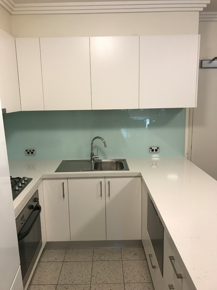 Photo of a mid-sized contemporary u-shaped kitchen in Sydney with a single-bowl sink, flat-panel cabinets, white cabinets, blue splashback, glass sheet splashback and black appliances.