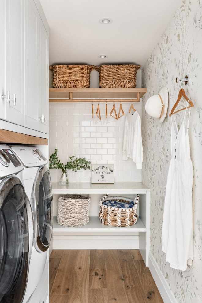 Inspiration for a transitional single-wall dedicated laundry room in Vancouver with recessed-panel cabinets, white cabinets, medium hardwood floors, a side-by-side washer and dryer, brown floor, white benchtop and wallpaper.