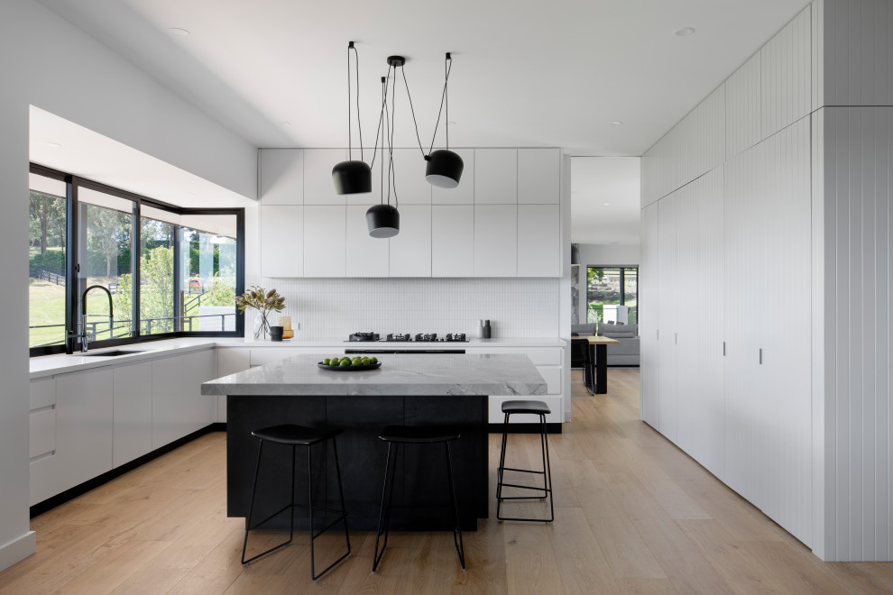 Photo of an expansive contemporary l-shaped eat-in kitchen in Melbourne with a drop-in sink, flat-panel cabinets, white cabinets, quartz benchtops, white splashback, ceramic splashback, black appliances, light hardwood floors, with island, brown floor and white benchtop.