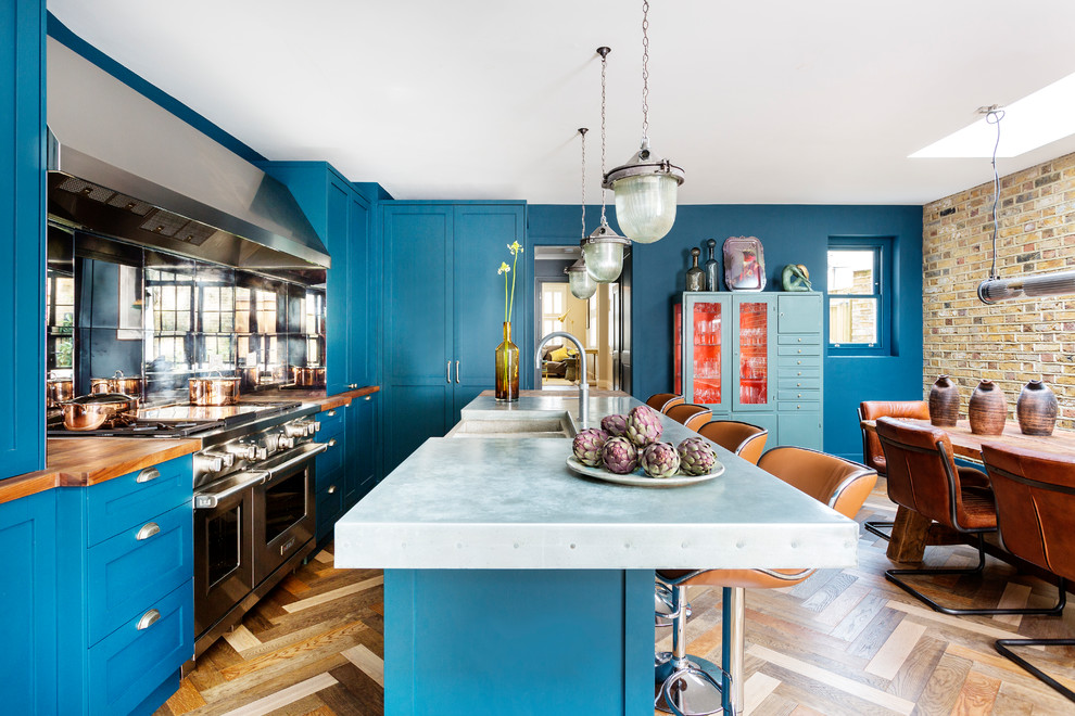 This is an example of a mid-sized eclectic l-shaped eat-in kitchen in London with a farmhouse sink, shaker cabinets, blue cabinets, zinc benchtops, mirror splashback, stainless steel appliances, with island, grey benchtop, medium hardwood floors and brown floor.