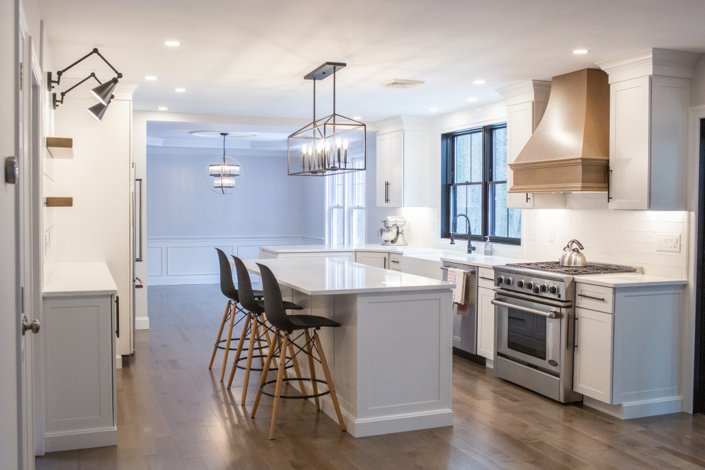 Photo of a large transitional kitchen in Boston with a farmhouse sink, recessed-panel cabinets, white cabinets, quartz benchtops, white splashback, subway tile splashback, stainless steel appliances, medium hardwood floors, with island and white benchtop.