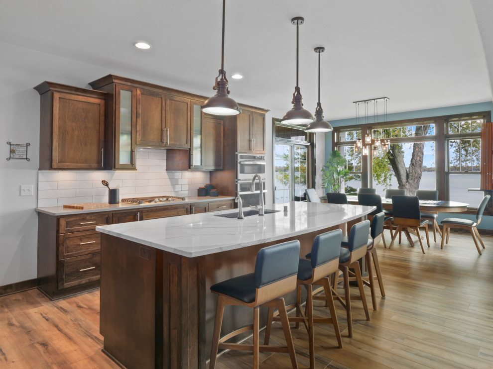Photo of an expansive country l-shaped eat-in kitchen in Milwaukee with an undermount sink, recessed-panel cabinets, dark wood cabinets, quartz benchtops, white splashback, subway tile splashback, stainless steel appliances, vinyl floors, with island, brown floor, white benchtop and vaulted.
