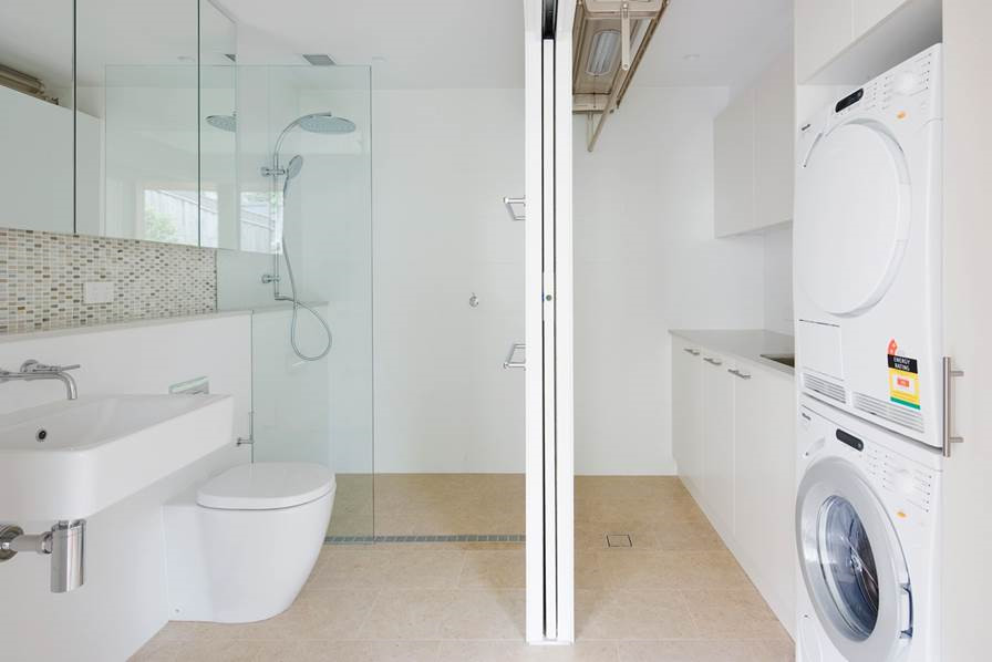 Photo of a mid-sized modern single-wall utility room in Sydney with an undermount sink, flat-panel cabinets, white cabinets, quartz benchtops, white walls, travertine floors and a stacked washer and dryer.