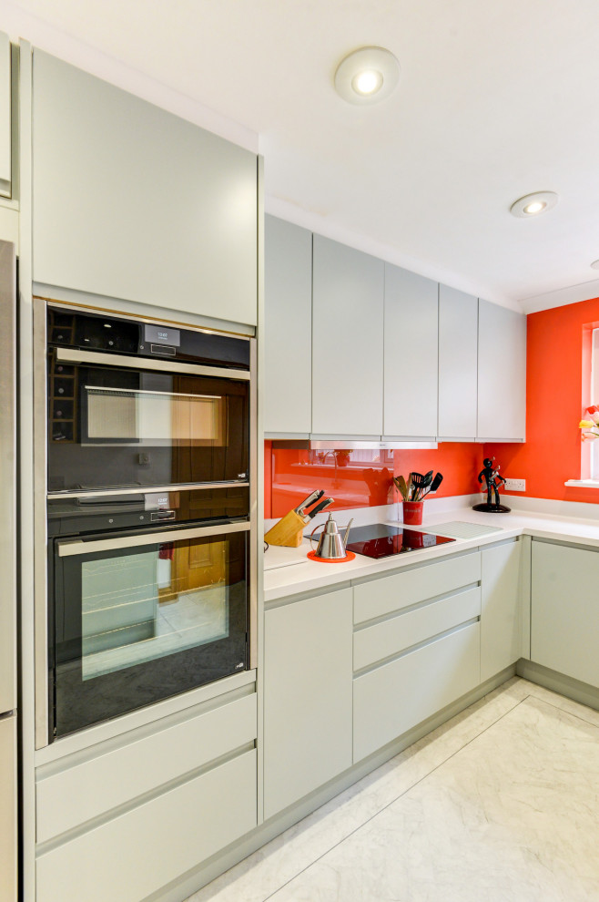 Photo of a medium sized beach style grey and white l-shaped enclosed kitchen in Sussex with a built-in sink, flat-panel cabinets, green cabinets, composite countertops, orange splashback, glass sheet splashback, black appliances, lino flooring, a breakfast bar, white floors, white worktops and feature lighting.