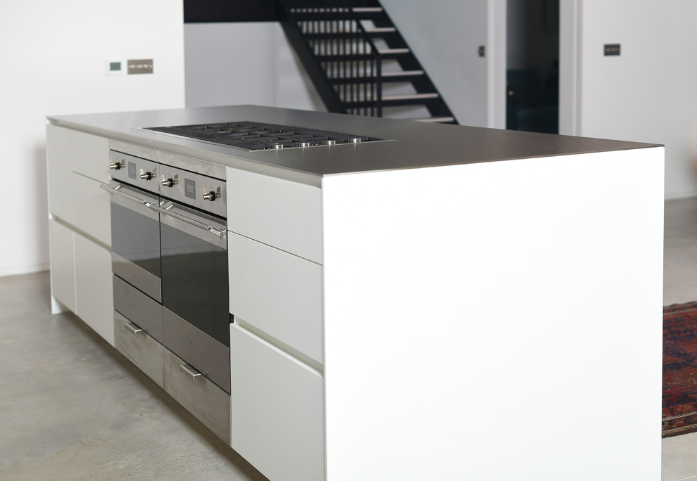 Photo of a contemporary kitchen in London with an integrated sink, flat-panel cabinets, white cabinets, stainless steel worktops, metallic splashback, stainless steel appliances, concrete flooring, an island and grey floors.