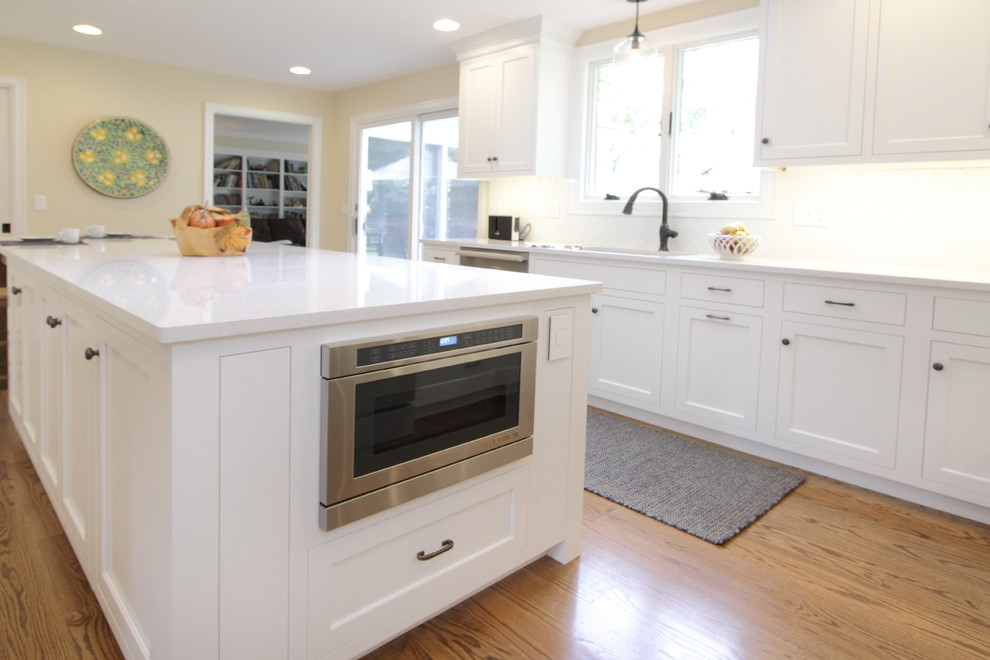 Example of a large transitional l-shaped medium tone wood floor and brown floor enclosed kitchen design in Philadelphia with an undermount sink, shaker cabinets, white cabinets, quartz countertops, white backsplash, ceramic backsplash, stainless steel appliances and an island