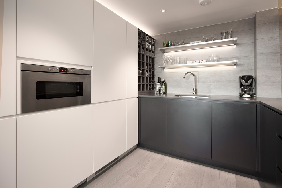 Photo of a small industrial u-shaped open plan kitchen in London with a drop-in sink, flat-panel cabinets, grey cabinets, quartz benchtops, grey splashback, ceramic splashback, panelled appliances, light hardwood floors and a peninsula.
