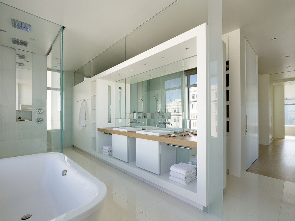 Photo of a large contemporary master bathroom in San Francisco with flat-panel cabinets, white cabinets, a freestanding tub, beige tile, stone tile, white walls, porcelain floors, a drop-in sink and wood benchtops.