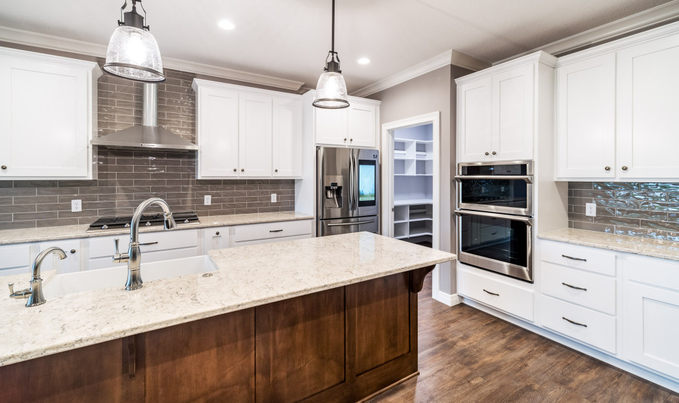 Inspiration for a transitional vinyl floor kitchen remodel with a farmhouse sink, shaker cabinets, white cabinets, quartz countertops, gray backsplash, subway tile backsplash, stainless steel appliances, an island and gray countertops
