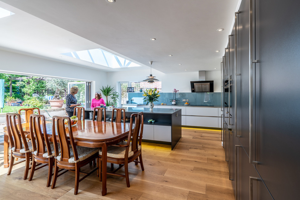 This is an example of an expansive modern open plan kitchen in Hampshire with an undermount sink, flat-panel cabinets, grey cabinets, quartz benchtops, blue splashback, glass sheet splashback, stainless steel appliances, medium hardwood floors, with island and grey benchtop.
