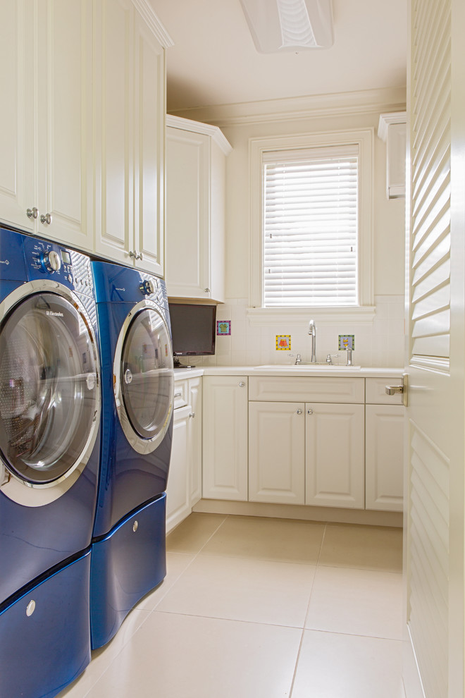 Inspiration for a large traditional l-shaped dedicated laundry room in Miami with a drop-in sink, raised-panel cabinets, white cabinets, solid surface benchtops, white walls, porcelain floors and a side-by-side washer and dryer.