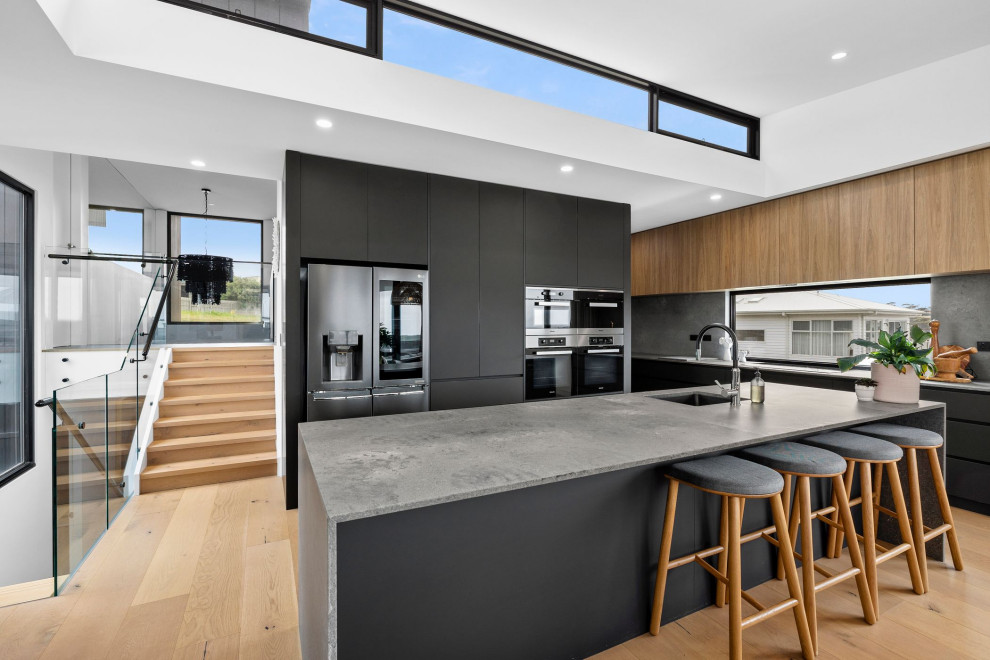 Design ideas for an expansive contemporary kitchen pantry in Geelong with a double-bowl sink, flat-panel cabinets, black cabinets, quartz benchtops, grey splashback, engineered quartz splashback, black appliances, light hardwood floors, with island and grey benchtop.