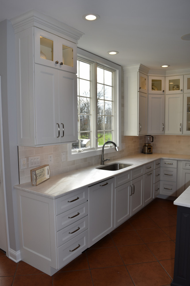 Klassisk inredning av ett stort vit vitt kök, med en nedsänkt diskho, släta luckor, vita skåp, bänkskiva i kvarts, vitt stänkskydd, stänkskydd i tunnelbanekakel, rostfria vitvaror, klinkergolv i terrakotta, en köksö och brunt golv