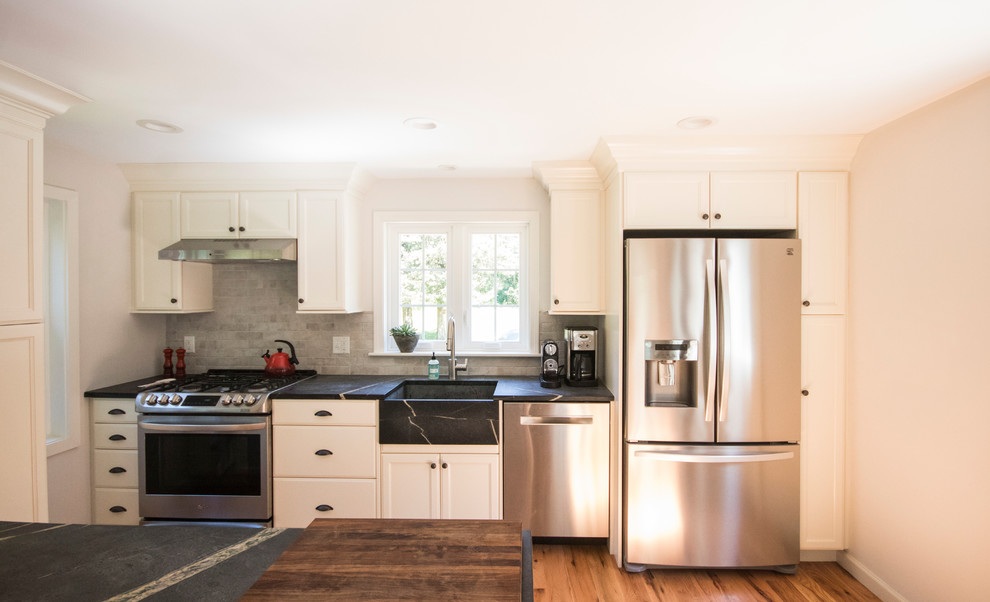 Expansive country l-shaped eat-in kitchen in Providence with a farmhouse sink, recessed-panel cabinets, white cabinets, soapstone benchtops, white splashback, porcelain splashback, stainless steel appliances, light hardwood floors, with island and beige floor.
