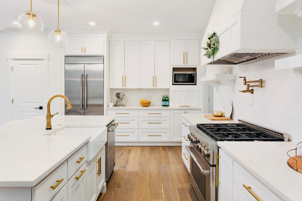 Design ideas for a mid-sized transitional u-shaped eat-in kitchen in Los Angeles with a farmhouse sink, shaker cabinets, white cabinets, quartz benchtops, white splashback, subway tile splashback, stainless steel appliances, medium hardwood floors, with island, brown floor and white benchtop.