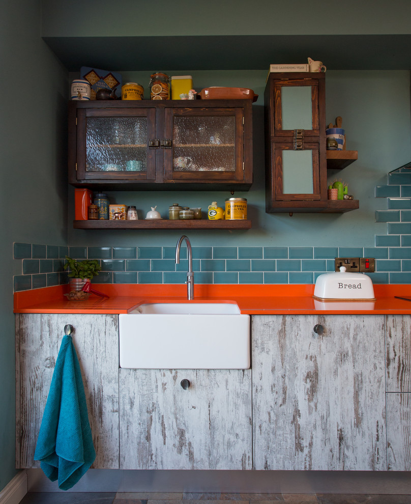 Photo of a mid-sized eclectic single-wall open plan kitchen in Other with a farmhouse sink, flat-panel cabinets, distressed cabinets, solid surface benchtops, blue splashback, ceramic splashback, stainless steel appliances, porcelain floors, with island and multi-coloured floor.