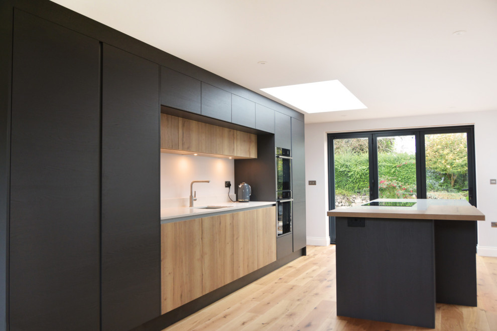 This is an example of a mid-sized contemporary galley open plan kitchen in Cornwall with an integrated sink, flat-panel cabinets, black cabinets, solid surface benchtops, black appliances, medium hardwood floors, with island and white benchtop.