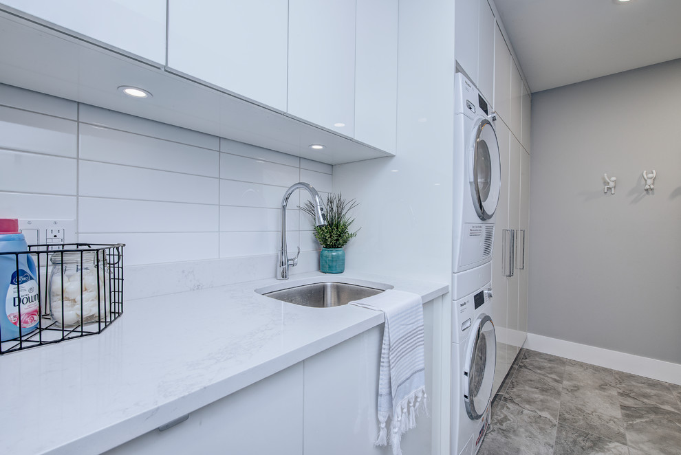 Inspiration for a mid-sized contemporary single-wall dedicated laundry room in Calgary with an undermount sink, flat-panel cabinets, white cabinets, quartz benchtops, grey walls, limestone floors, a stacked washer and dryer, grey floor and white benchtop.