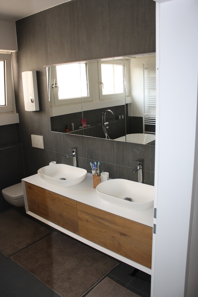 Photo of a contemporary bathroom in Essen with flat-panel cabinets, brown cabinets, a freestanding tub, a curbless shower, a wall-mount toilet, black tile, ceramic tile, white walls, a wall-mount sink, solid surface benchtops, black floor, an open shower and white benchtops.