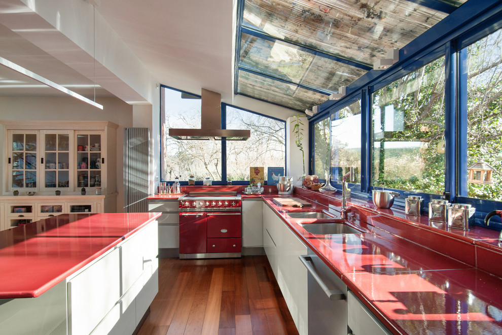 This is an example of a large midcentury galley eat-in kitchen in Marseille with a double-bowl sink, flat-panel cabinets, grey cabinets, solid surface benchtops, red splashback, panelled appliances, medium hardwood floors, with island, brown floor and red benchtop.
