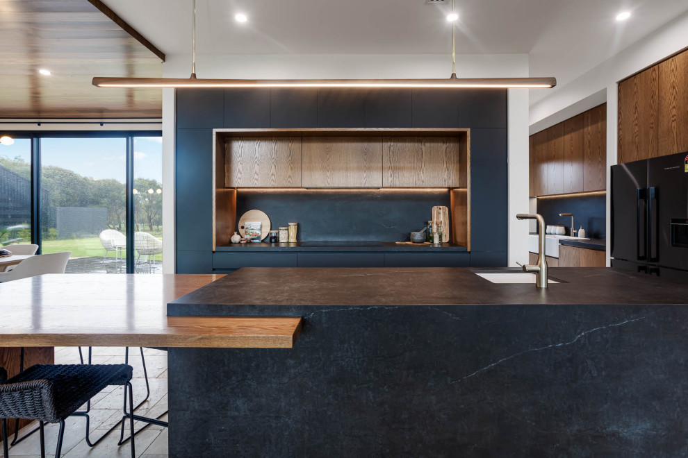 Inspiration for a large industrial open plan kitchen in Adelaide with a farmhouse sink, black cabinets, quartz benchtops, black splashback, stone slab splashback, black appliances, travertine floors, with island, beige floor and black benchtop.