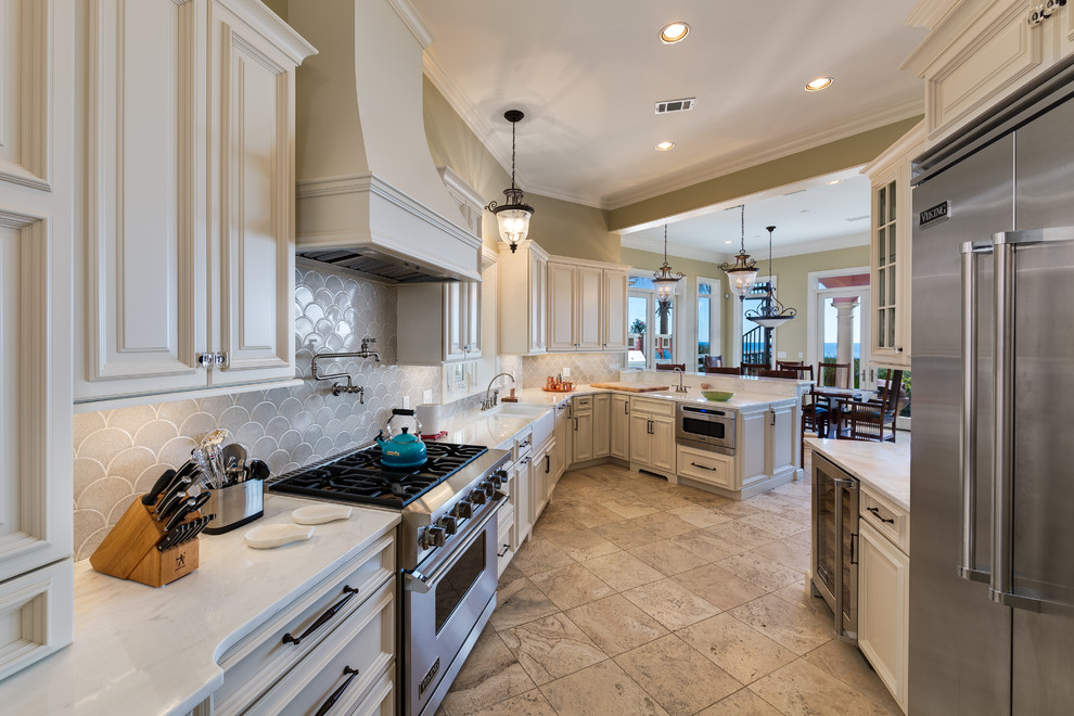 This is an example of a large traditional galley eat-in kitchen in Miami with raised-panel cabinets, white cabinets, marble benchtops, a peninsula, a farmhouse sink, white splashback, ceramic splashback, stainless steel appliances and travertine floors.
