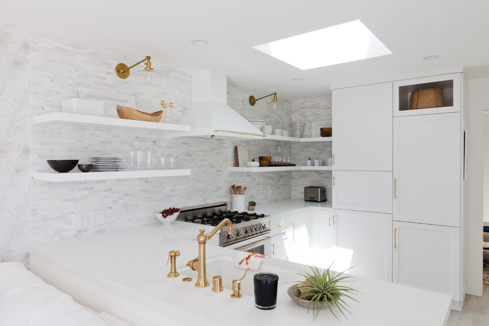 Inspiration for a mid-sized beach style u-shaped open plan kitchen in Los Angeles with recessed-panel cabinets, white cabinets, marble benchtops, white splashback, porcelain splashback, white appliances, light hardwood floors, no island and a single-bowl sink.