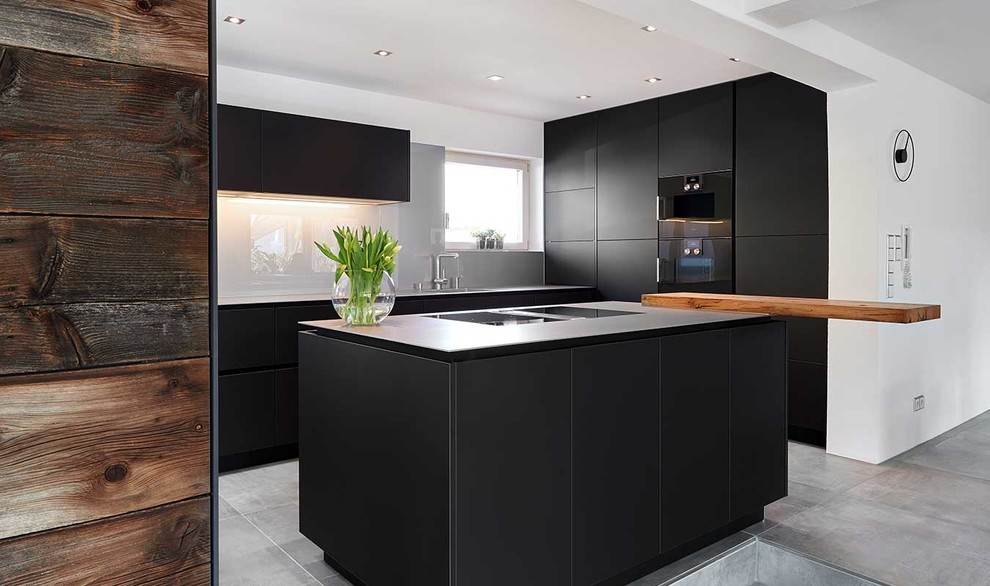 Photo of a mid-sized contemporary l-shaped open plan kitchen in Stuttgart with a drop-in sink, flat-panel cabinets, black cabinets, stainless steel benchtops, white splashback, glass sheet splashback, black appliances, concrete floors, with island and grey floor.