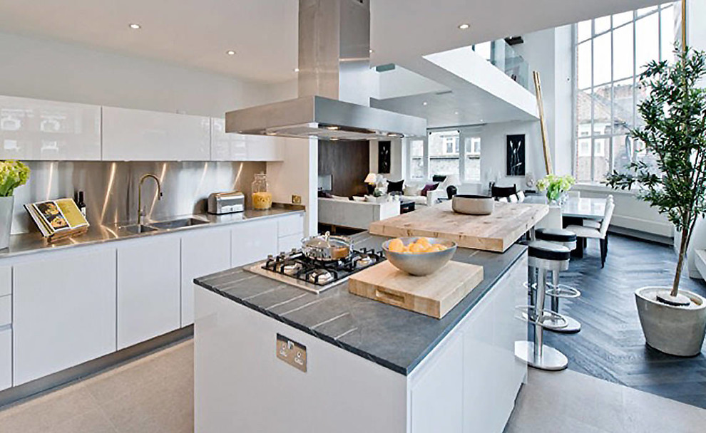 Large contemporary single-wall open plan kitchen in London with an integrated sink, recessed-panel cabinets, white cabinets, granite worktops, metallic splashback, metal splashback, integrated appliances, cement flooring, an island and grey floors.