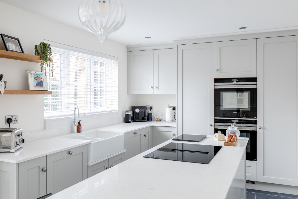 Mid-sized traditional l-shaped open plan kitchen in Hertfordshire with a farmhouse sink, shaker cabinets, grey cabinets, quartzite benchtops, glass sheet splashback, with island and white benchtop.
