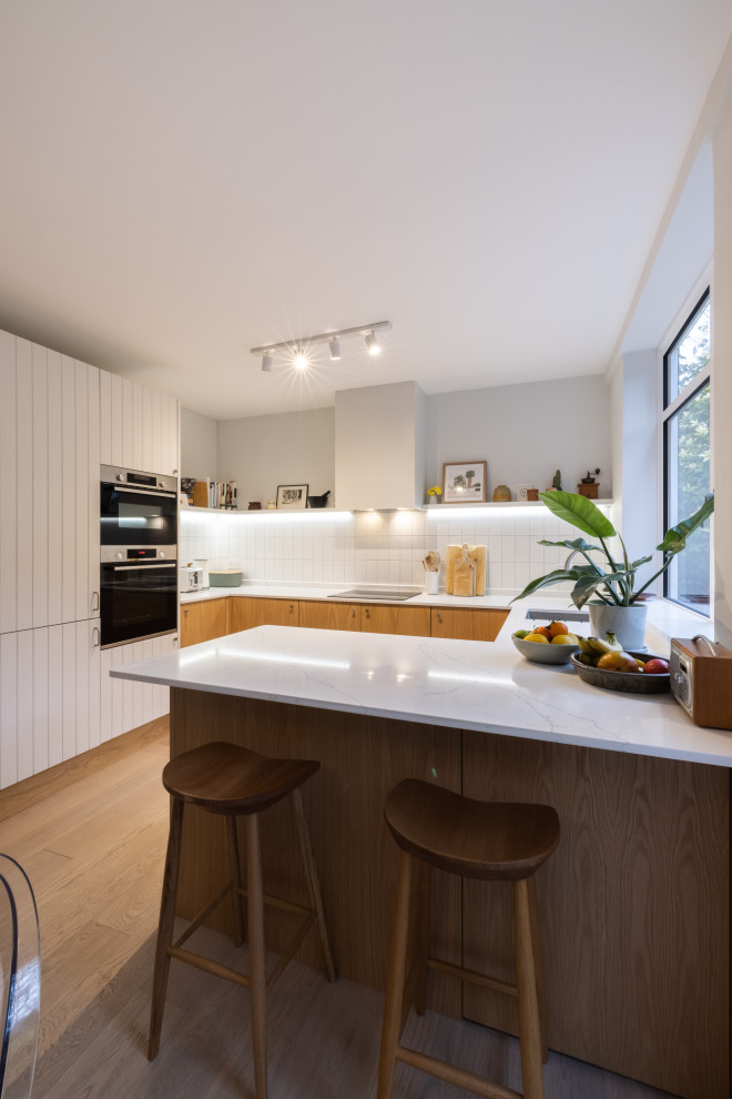 Medium sized contemporary u-shaped kitchen/diner in London with a submerged sink, louvered cabinets, light wood cabinets, quartz worktops, white splashback, ceramic splashback, stainless steel appliances, light hardwood flooring, a breakfast bar, brown floors and white worktops.