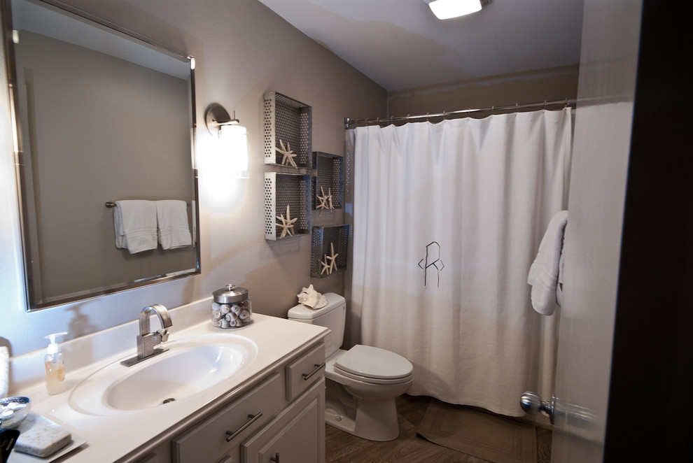 Mid-sized beach style bathroom in Houston with raised-panel cabinets, white cabinets, solid surface benchtops, a shower/bathtub combo, grey walls, light hardwood floors, an alcove tub, a two-piece toilet and an integrated sink.
