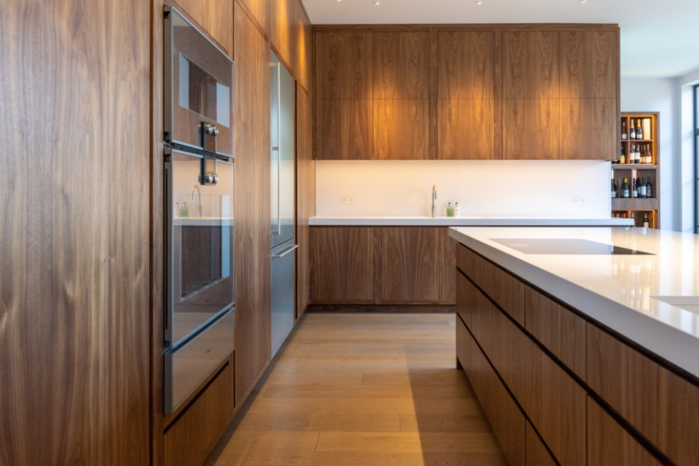 Large midcentury l-shaped eat-in kitchen in London with an integrated sink, flat-panel cabinets, medium wood cabinets, quartzite benchtops, white splashback, stone slab splashback, stainless steel appliances, medium hardwood floors, with island, brown floor and white benchtop.