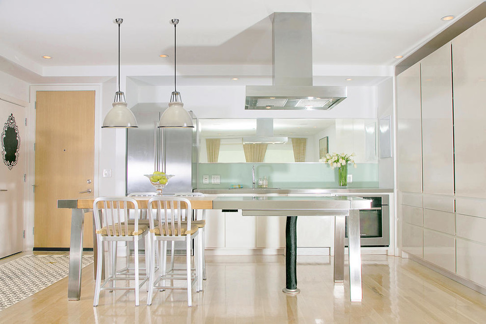 Photo of a contemporary galley kitchen in Chicago with flat-panel cabinets, white cabinets, stainless steel benchtops, green splashback, glass sheet splashback, stainless steel appliances, light hardwood floors and with island.